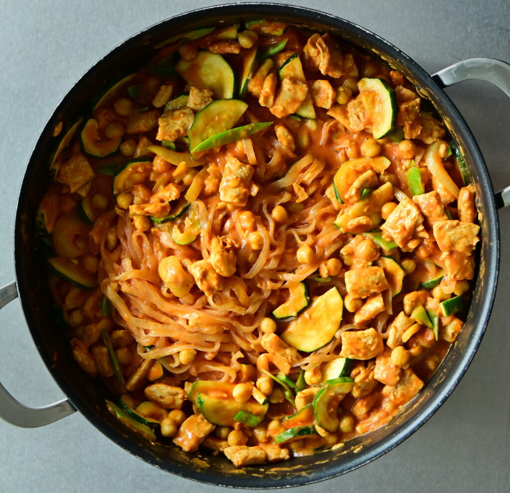 A wide, deep skillet filled with rosé noodles, chickpeas, zucchini, and vegan protein chunks on a metal countertop.