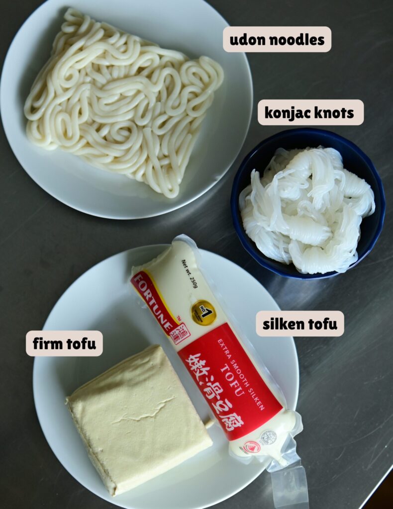 White plates with firm tofu and silken tofu, a white plate with udon noodles, and a blue bowl of konjac noodles on a metal surface.