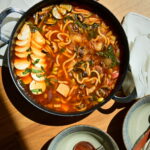 close up to a vegan Korean Army Stew (Budae Jjigae) in a shallow pot on a white tablecloth, with owls, chopsticks and spoons