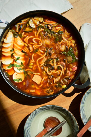 close up to a vegan Korean Army Stew (Budae Jjigae) in a shallow pot on a white tablecloth, with owls, chopsticks and spoons