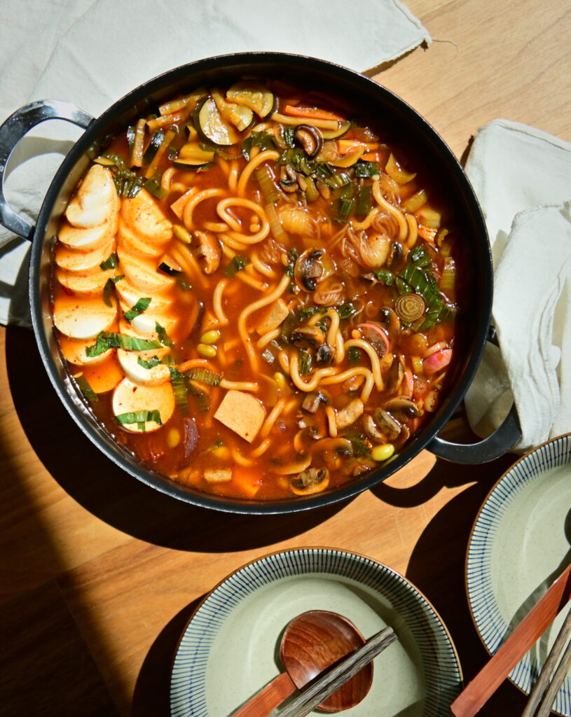 close up to a vegan Korean Army Stew (Budae Jjigae) in a shallow pot on a white tablecloth, with owls, chopsticks and spoons