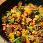 A wok filled with colorful stir-fried vegetables and seitan sausages on a wooden surface.