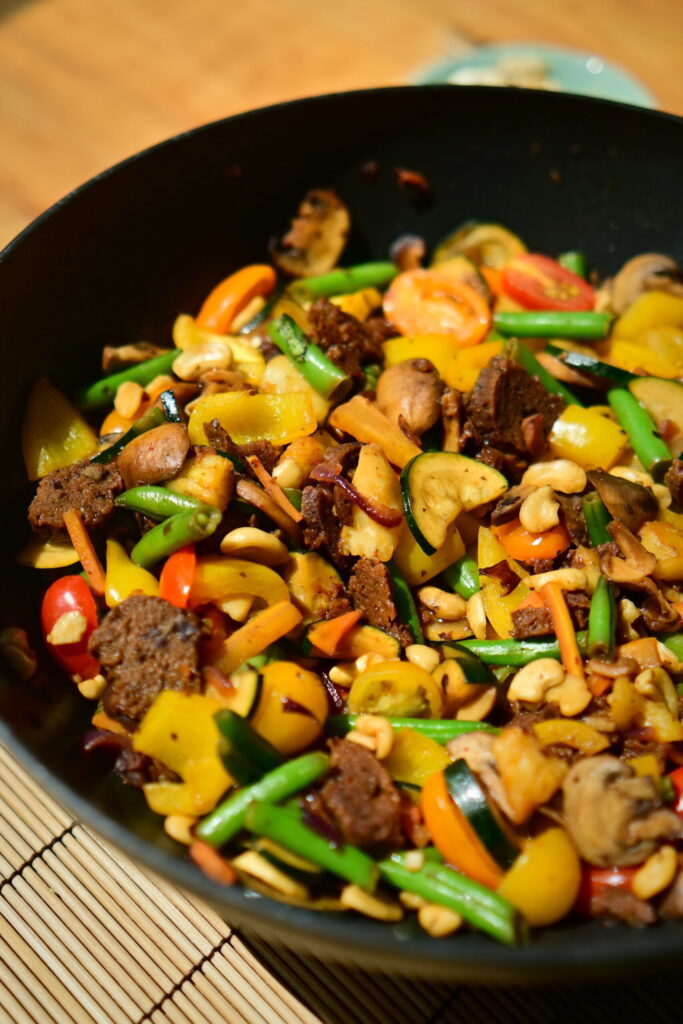 A wok filled with colorful stir-fried vegetables and seitan sausages on a wooden surface.