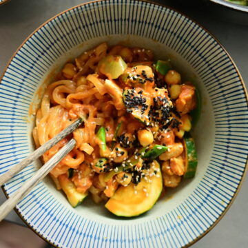 A striped deep bowl filled with rosé noodles, chickpeas, zucchini, and vegan protein chunks, with wooden chopsticks.
