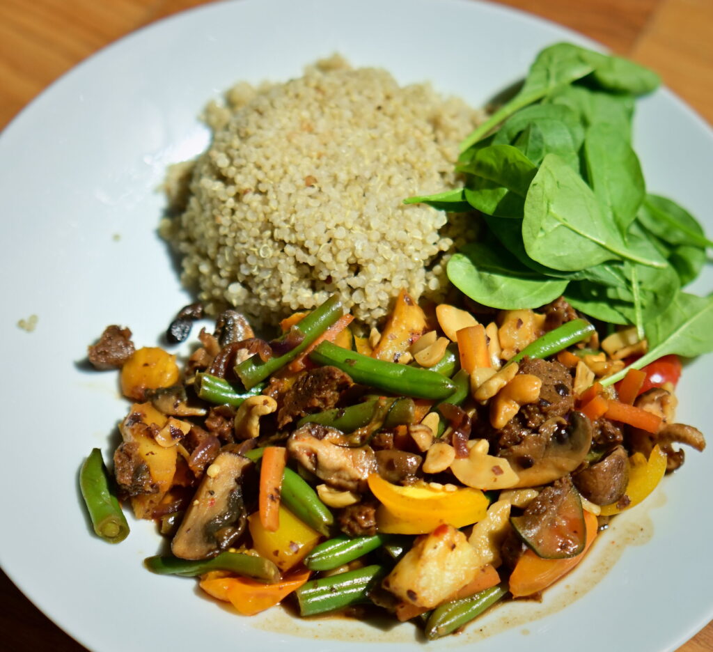 A white plate with colorful stir-fried vegetables and seitan sausages, served with quinoa and fresh spinach.