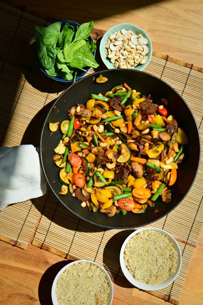 A wok filled with colorful stir-fried vegetables and seitan sausages on a wooden surface, with a small bowl of quinoa on the side.