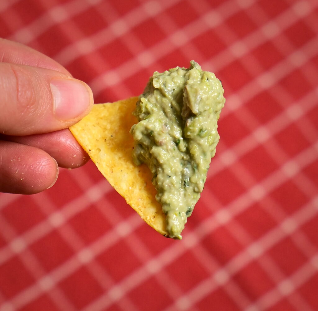 One nacho topped with pea-avocado spread, set against a red checkered tablecloth.