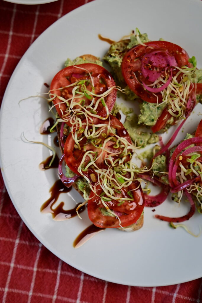 Avocado Pea Spread on sourdough toast topped with sprouts, Pickled red onion, served on a red checkered tablecloth.