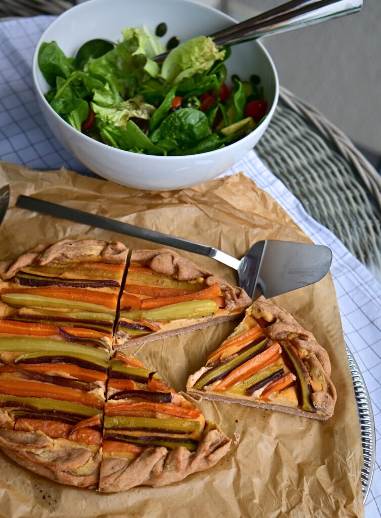 A slice of galette filled with colorful carrots, next to a serving spoon on parchment paper.