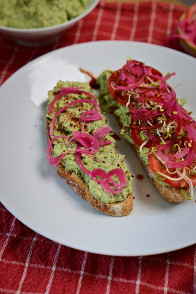 Avocado Pea Spread on sourdough toast topped with sprouts, Pickled red onion, served on a red checkered tablecloth.