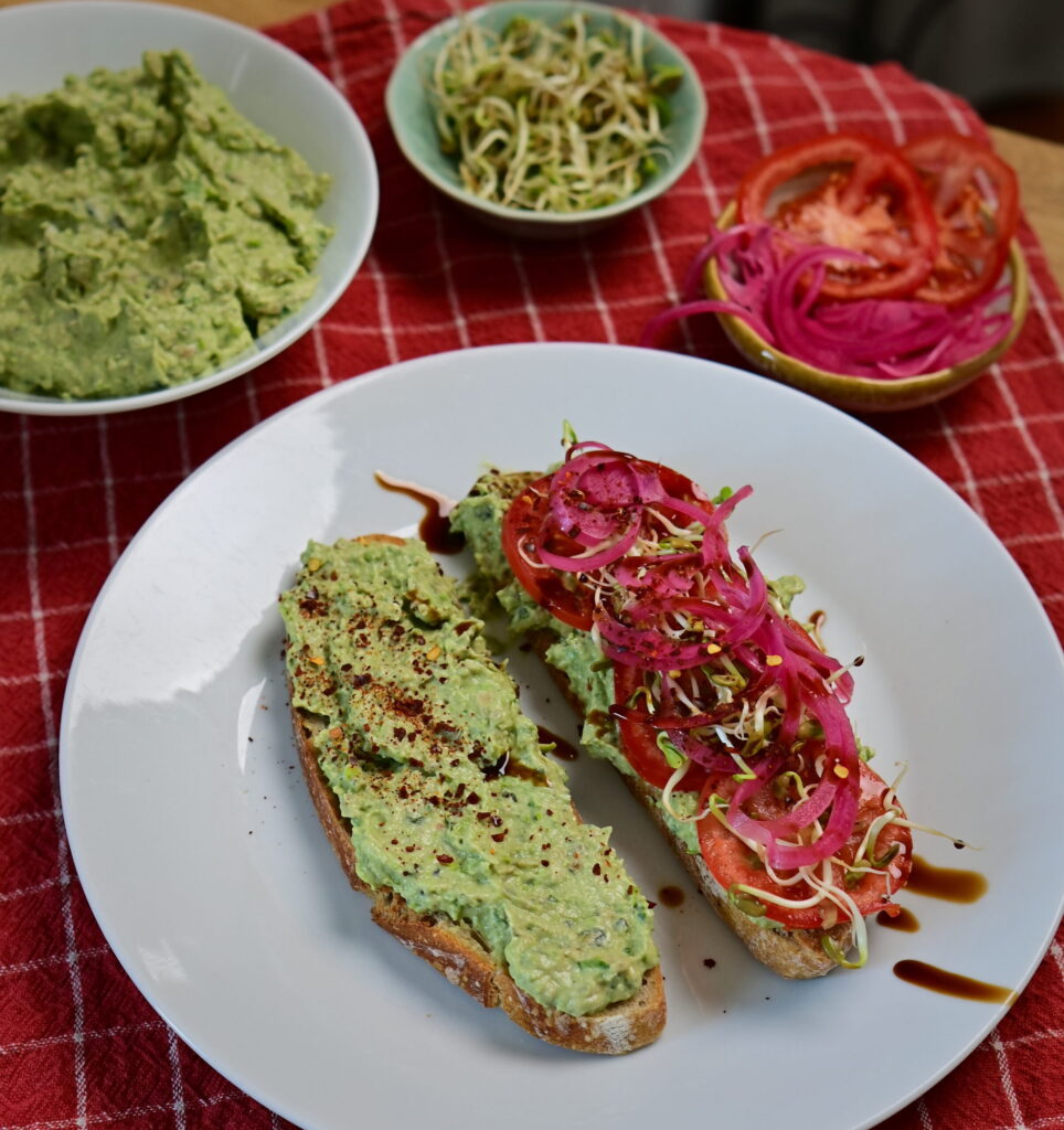 Avocado Pea Spread on sourdough toast topped with sprouts, Pickled red onion, served on a red checkered tablecloth.