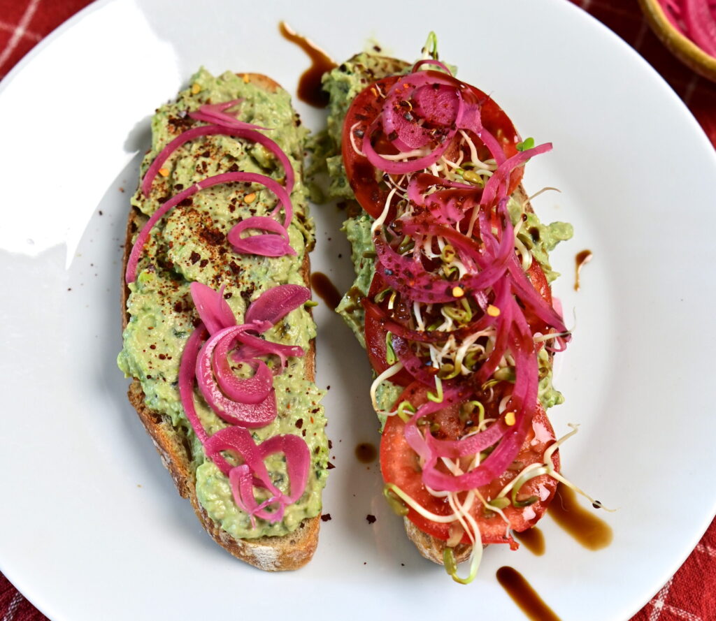Avocado Pea Spread on sourdough toast topped with sprouts, Pickled red onion, served on a red checkered tablecloth.