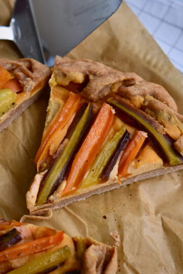 A slice of galette filled with colorful carrots, next to a serving spoon on parchment paper.