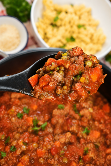 A spoon holding Veggie-Packed Vegan Mince over a pot and a bowl of pasta.