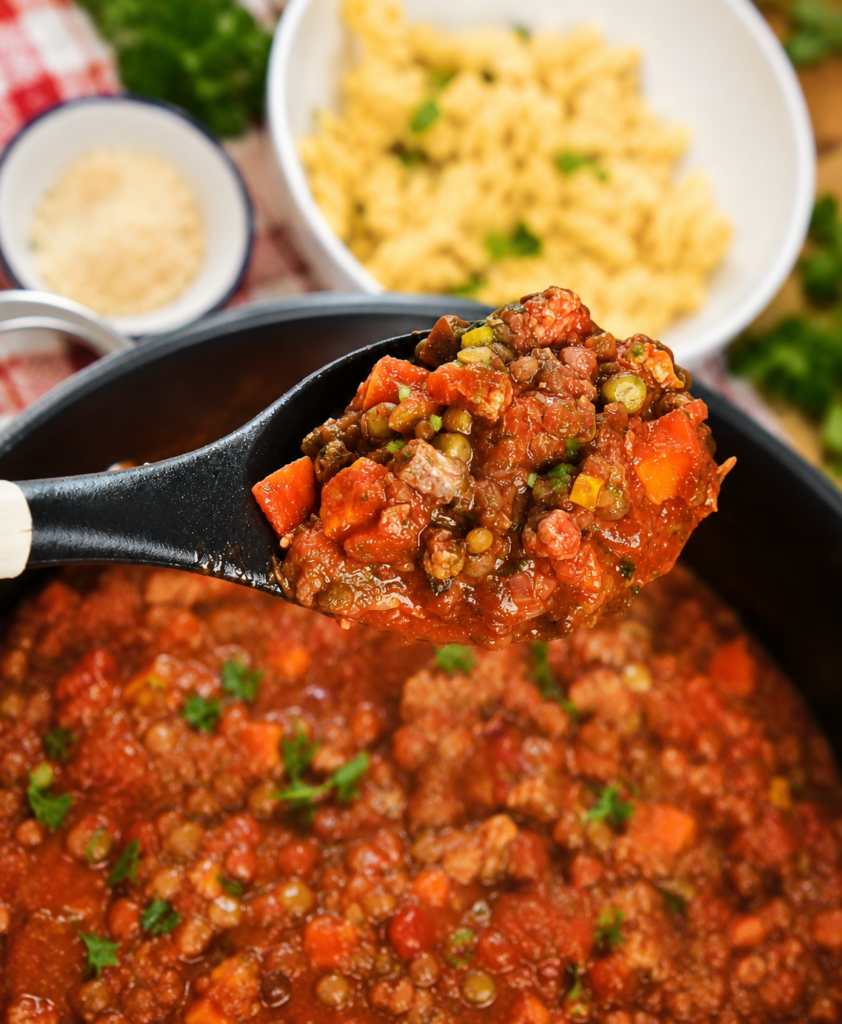 A spoon holding Veggie-Packed Vegan Mince over a pot and a bowl of pasta.